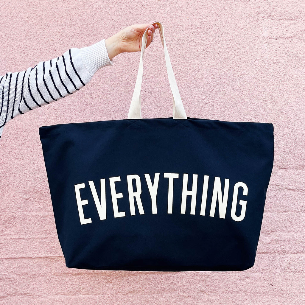 Navy tote bag with 'EVERYTHING' text held by a person against a pink background