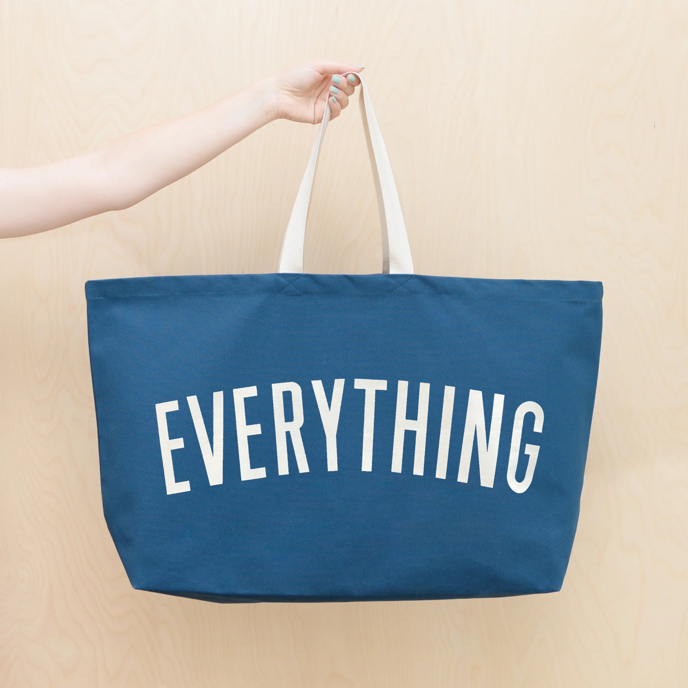 Blue tote bag with 'EVERYTHING' text held by a person against a beige background