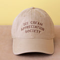 Beige cap with 'Ice Cream Appreciation Society' text on a beige surface with yellow background