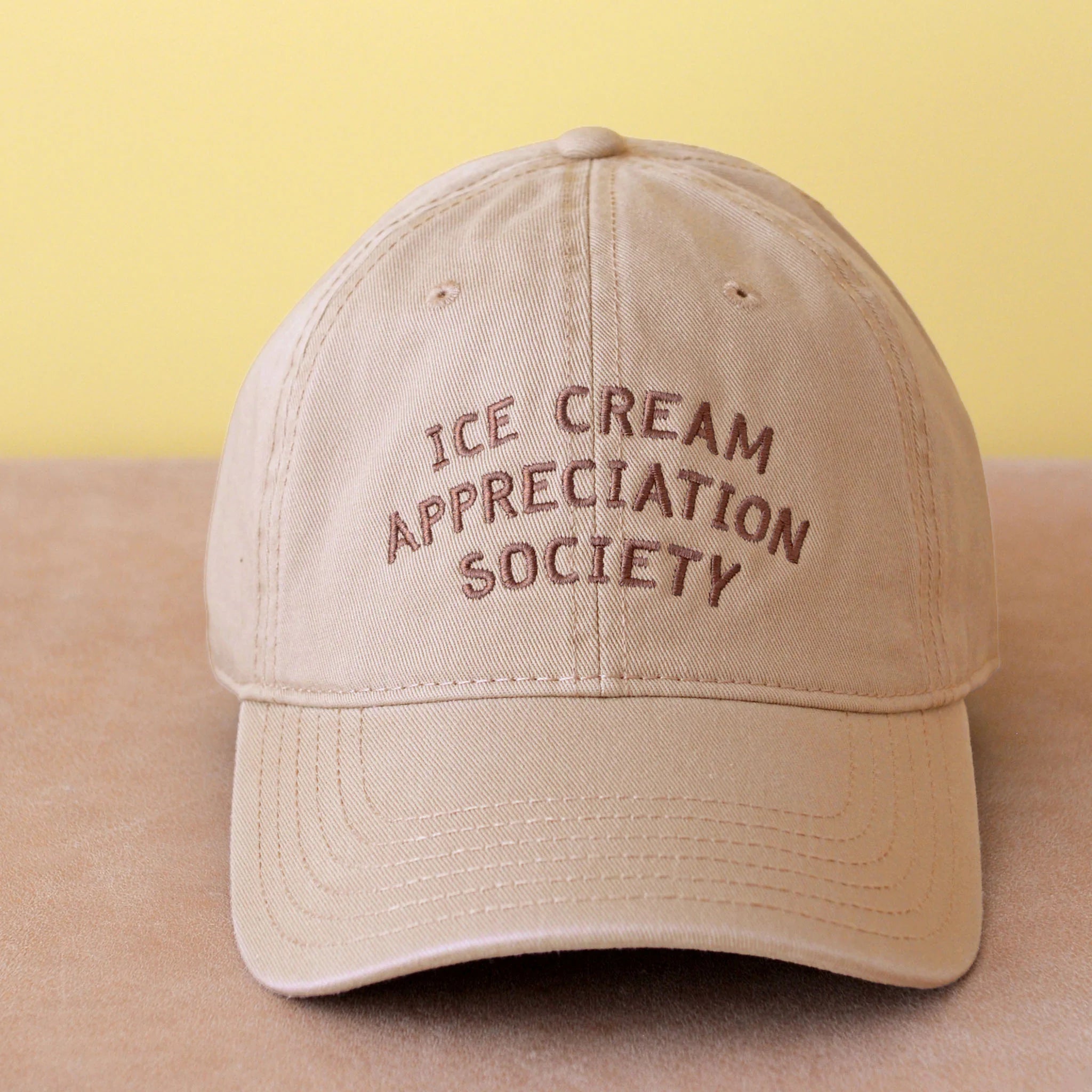 Beige cap with 'Ice Cream Appreciation Society' text on a beige surface with yellow background
